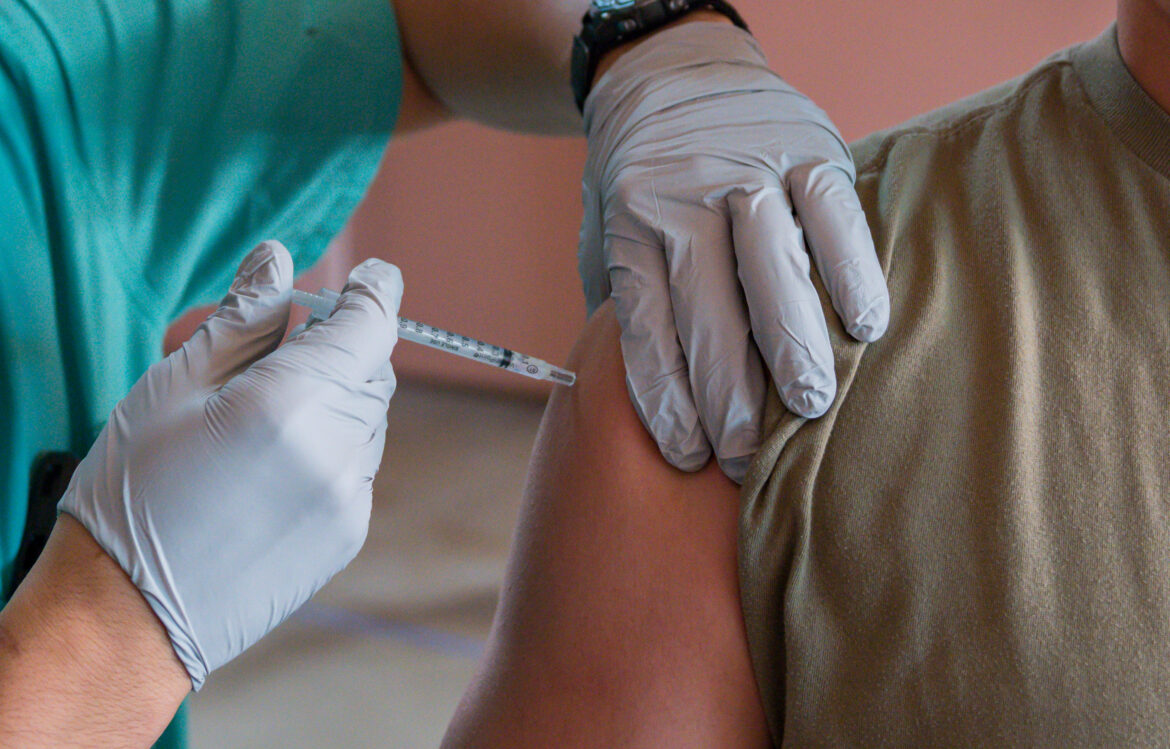 A gloved provider holds a needle to a person's shoulder in preparation for a vaccine injection.