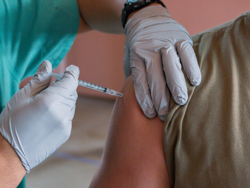 A gloved provider holds a needle to a person's shoulder in preparation for a vaccine injection.