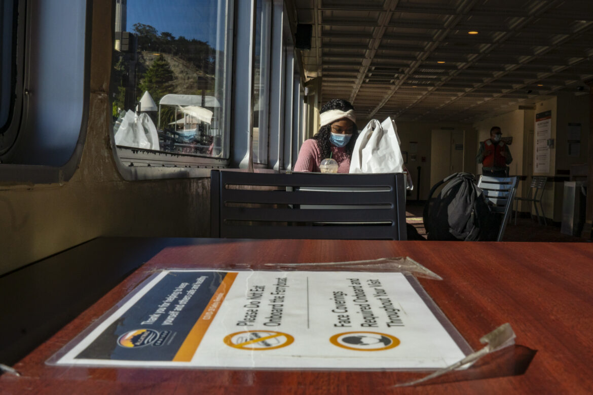 An Alcatraz employee is seated on a ferry to the island with a sign instructing COVID-19 protocol in the foreground.