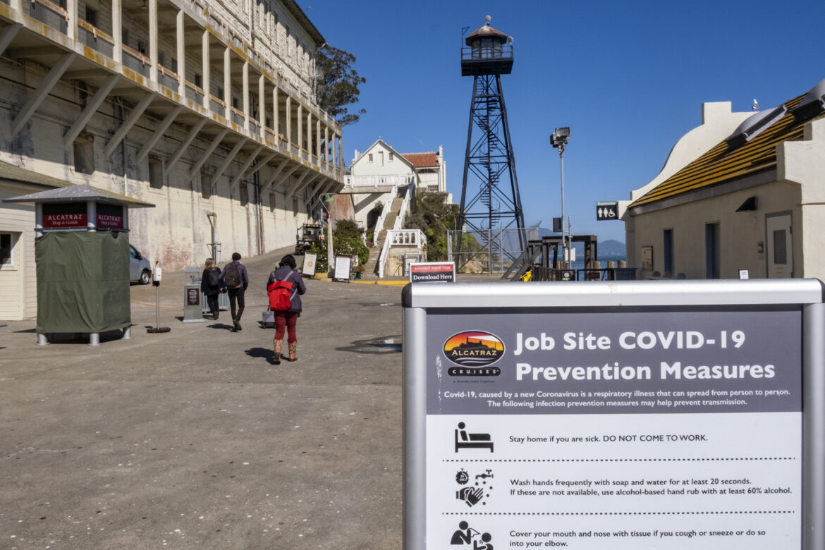 National Park employees arrive on Alcatraz, walking past historic buildings, to prepare the island for the day’s visitors. There is a COVID-19 protocol sign in the foreground and a water tower in the background.