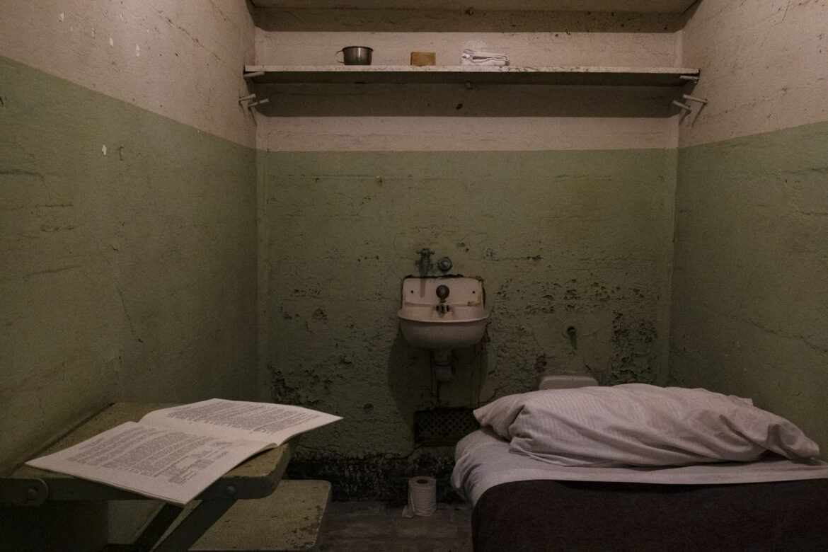 Inside a cell in the prison at Alcatraz with a sink, toilet, small desk, two shelves and a bed with a pillow and blankets.