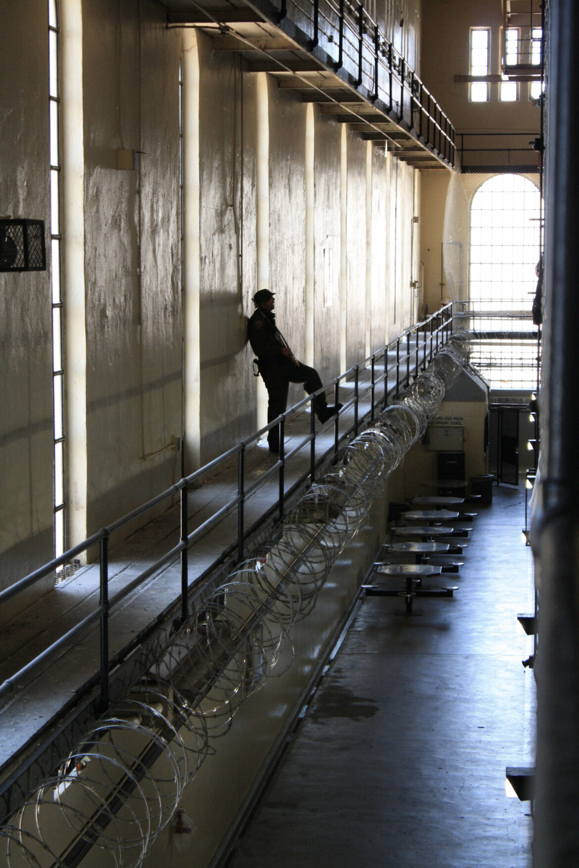 A prison guard stands inside a hall at San Quentin State Prison, his leg up on a railing fortified with razor wire, overlooking the ground floor.
