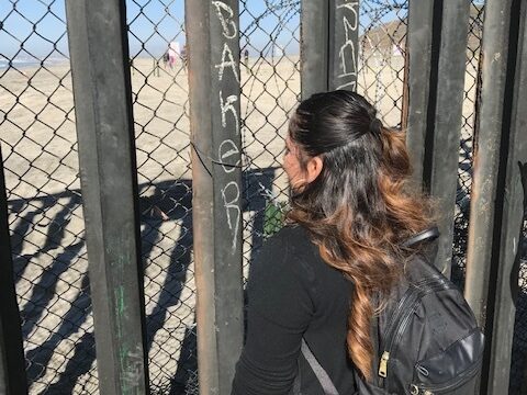 A woman with a backpack faces away from the viewer through a wire and post fence between the U.S.-Mexico border.