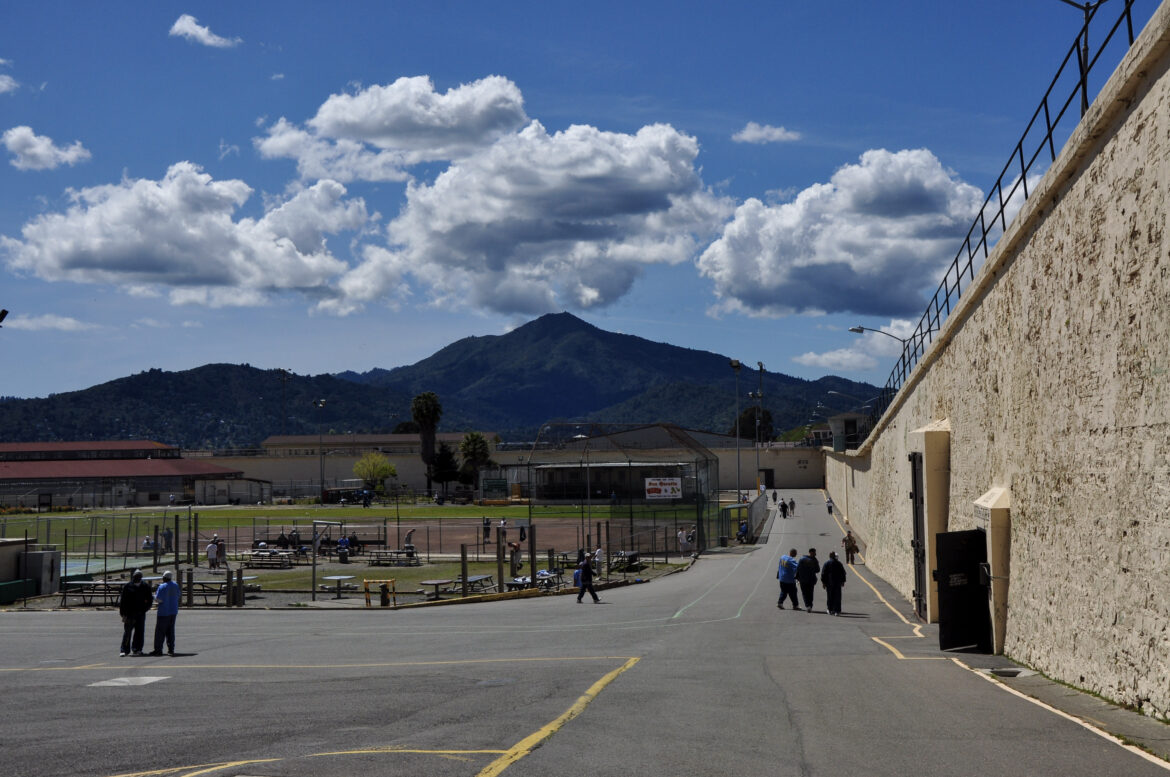 Several men gather just outside a building at San Quentin prison, with Mt. Tamalpais framed by dramatic clouds in the background. Four prisoners a tSan Quentin have tested positive for COVID-19 and their housing unit is under quarantine.