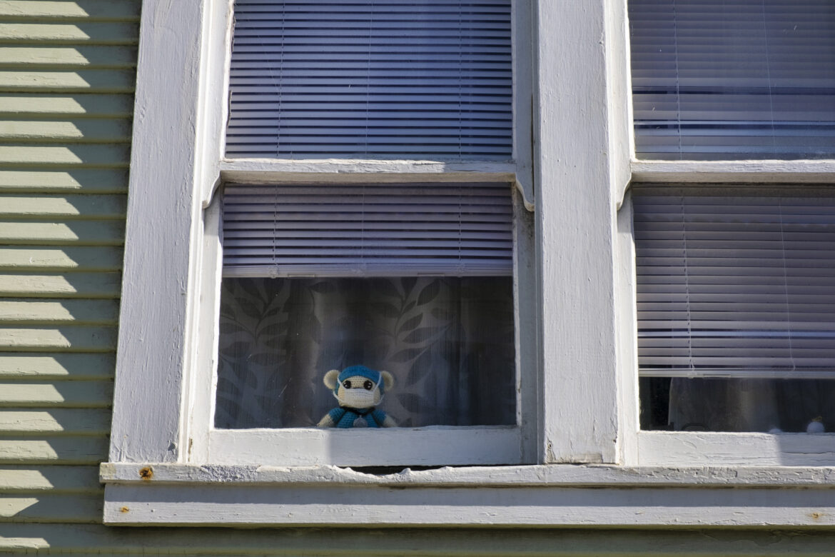 A crochet teddy bear peeks through the window of a family home in the Ingleside neighborhood. The teddy bear wears blue scrubs, a stethoscope and a mask.