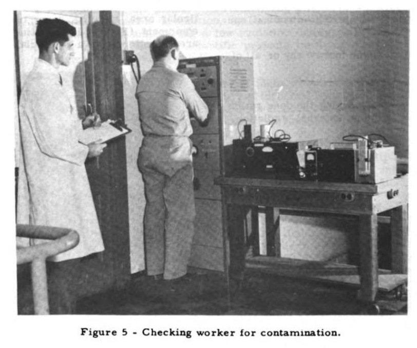 grainy black-and-white photo of two men in a laboratory, one standing in front of a large machine and another standing behind him in a white lab coat and holding a clipboard, with a caption saying