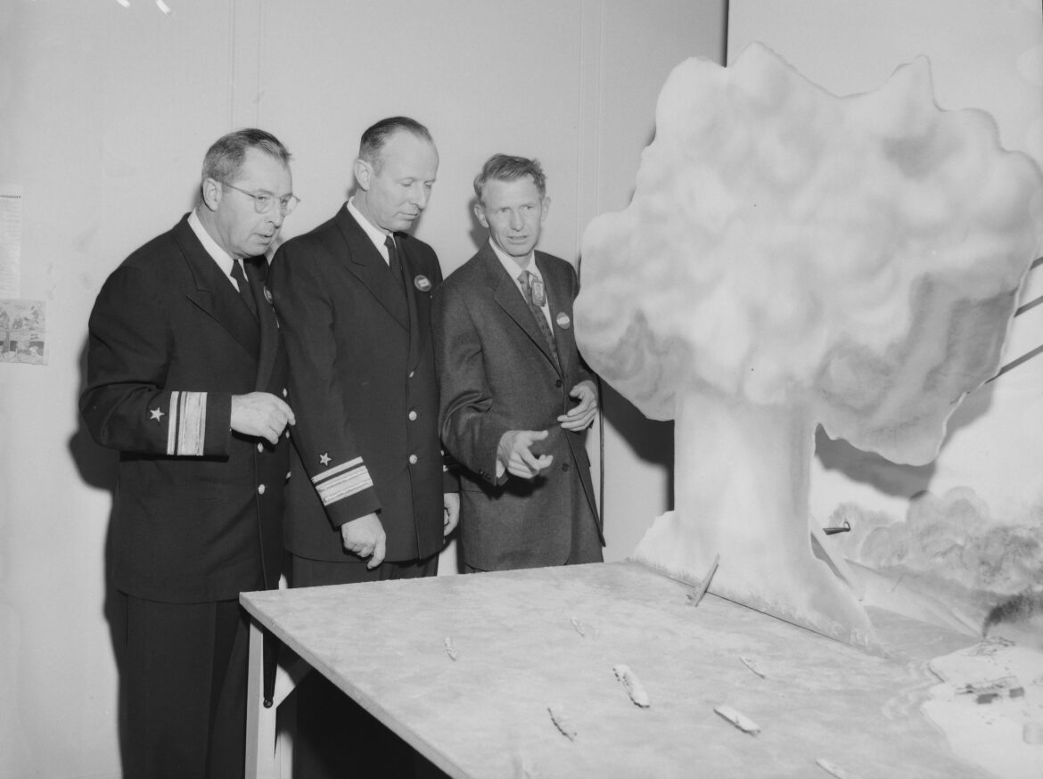 Three men in suits, two of them with military adornments, pose before a diorama of a mushroom cloud and ships at sea in a black-and-white photo