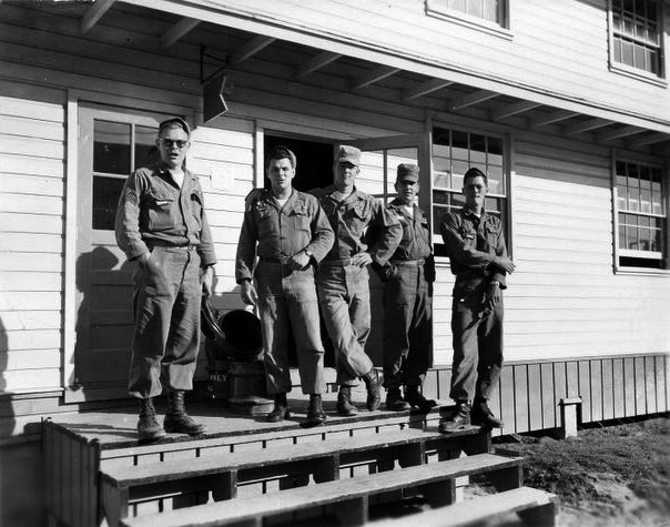 five men in uniform stand casually on a wooden porch in a black-and-white photo