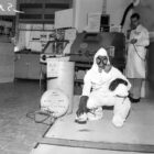 two men in a laboratory, one in a white lab coat and another in a gas mask and full-body hazard suit, posing in a black-and-white photo