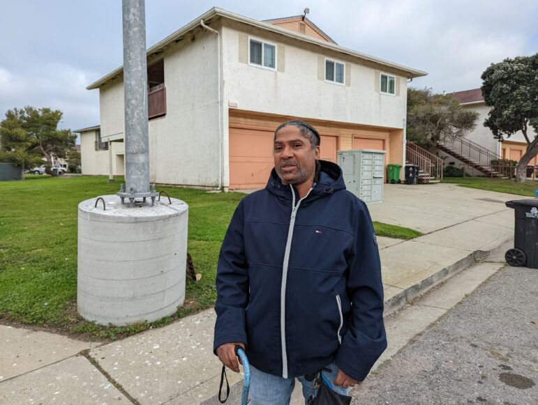 A Black man with a cane stands at an intersection in front of a grassy yard and a two-story house.Antonio Mixon moved to Treasure Island from San Francisco’s Tenderloin neighborhood six years ago after gaining custody of his daughter. His family has learned to adapt to the power outages, with an indoor backup generator and candles and flashlights in every room.