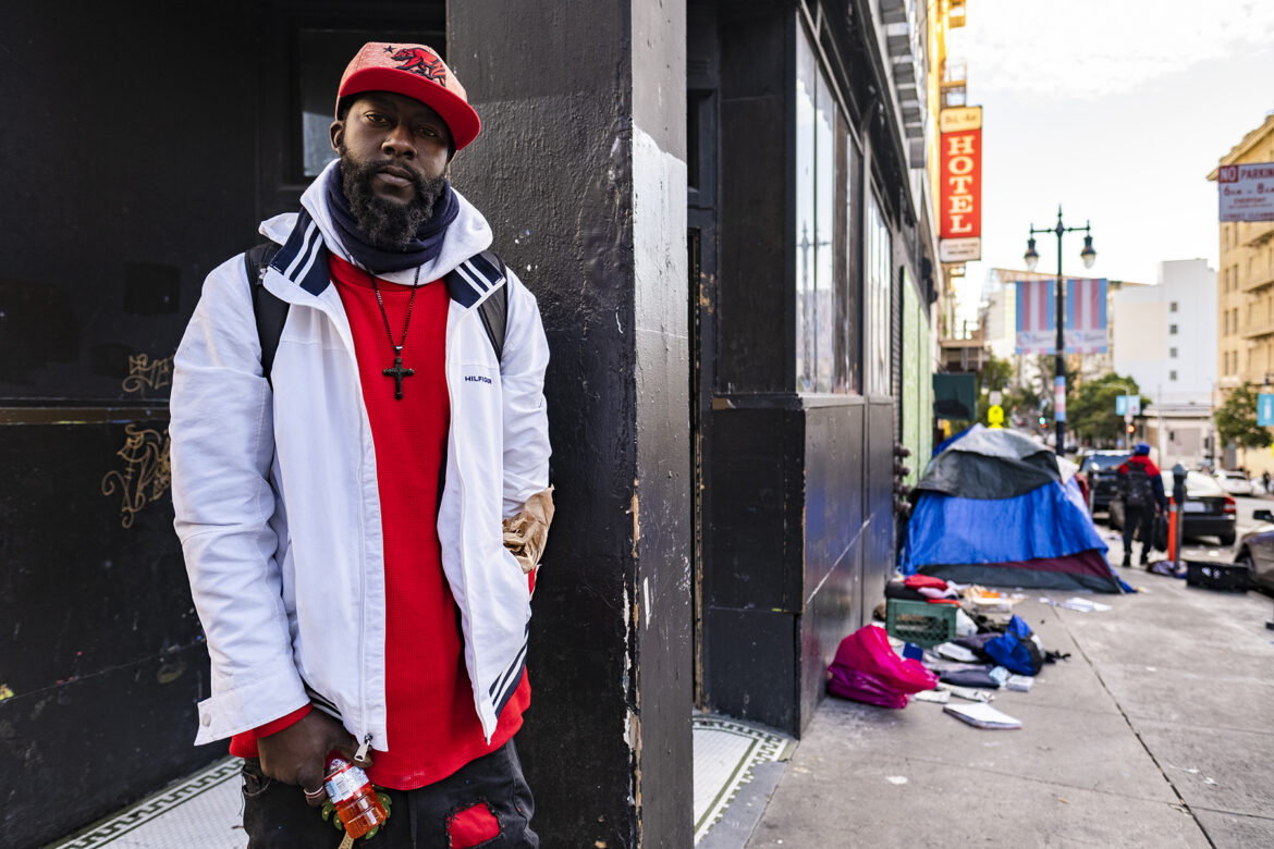 A young Black man stands on a San Francisco sidewalk. Down the sidewalk behind him sit tents and strewn clothing.