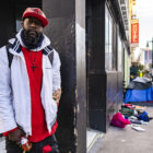 A young Black man stands on a San Francisco sidewalk. Down the sidewalk behind him sit tents and strewn clothing.