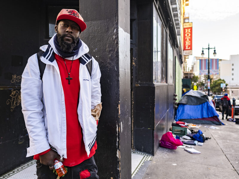 A young Black man stands on a San Francisco sidewalk. Down the sidewalk behind him sit tents and strewn clothing.