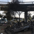 A tent stands to the left of a pile of charred debris under the charred bottom of an overpass.