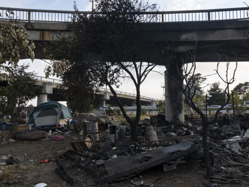 A tent stands to the left of a pile of charred debris under the charred bottom of an overpass.