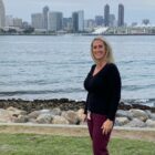 A white woman with blond hair stands on a lawn in front of the water, with a city skyline in the background.