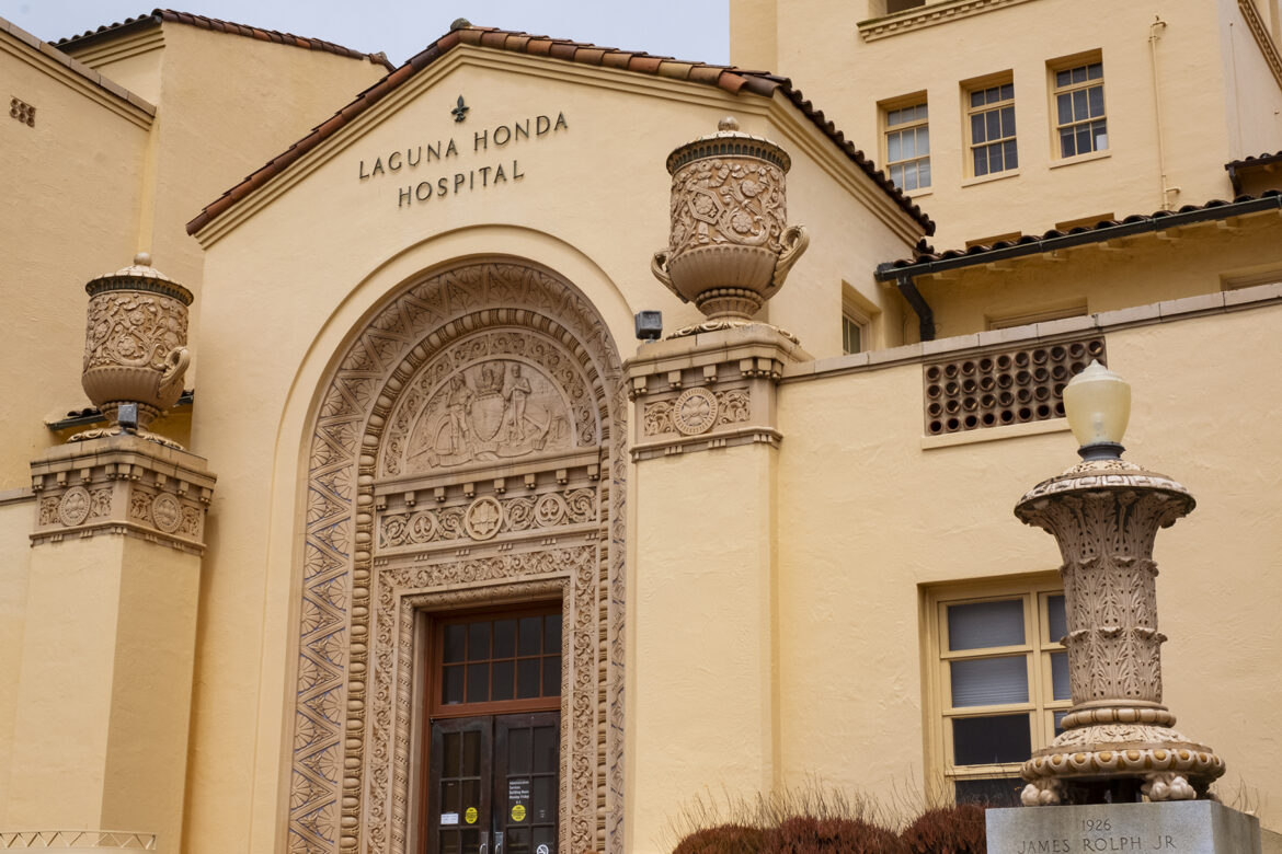 Two carved stone urns on pillars flank the arched entrance of Laguna Honda Hospital. The exterior walls are pale peach stucco topped by rred terra cotta roof tiles. The architecture is in the Spanish Revival style.