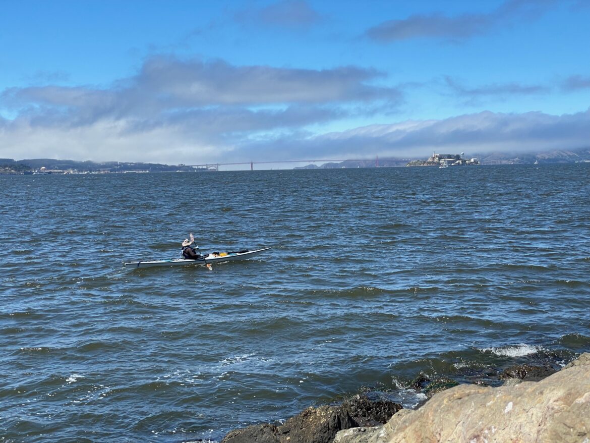 A kayaker paddles past Treasure Island as fog rolls in over the Presidio and Marin Headlands and across the Golden Gate Bridge into the San Francisco Bay.