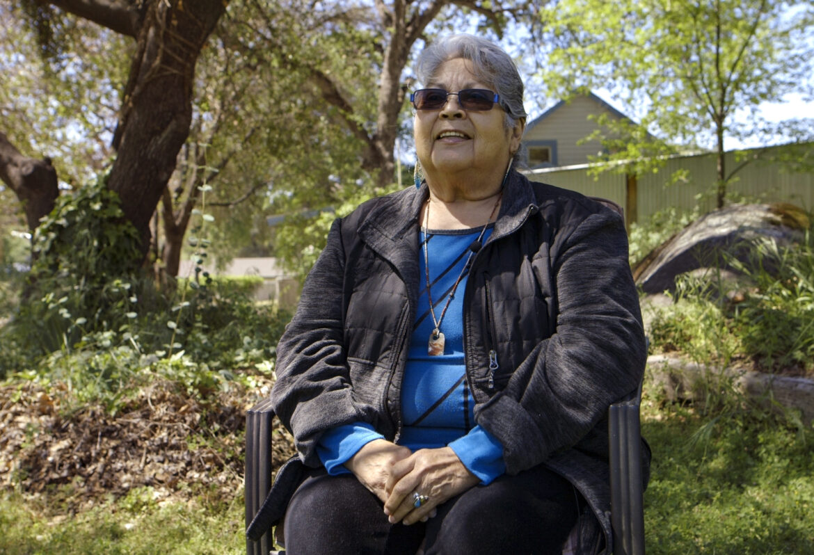 A Native American woman in a blue shirt and black jacket sits on a chair in a forested area near a house.