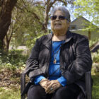 A Native American woman in a blue shirt and black jacket sits on a chair in a forested area near a house.