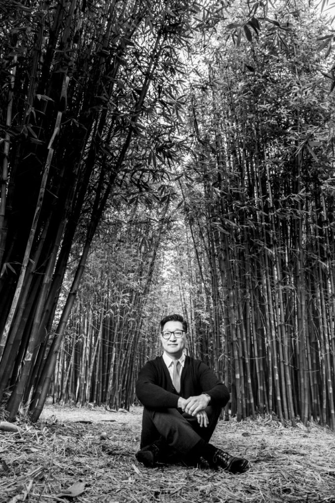 In this black-and-white photo, a man facing the camera sits outdoors on ground covered with dry leaves in front of a stand of tall, leafy bamboo.