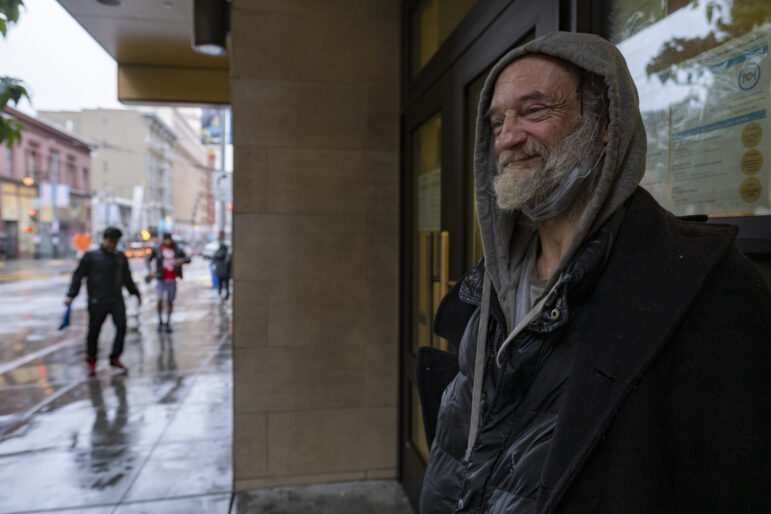 A man with a white beard in a grey hoodie and black puffy jacket stands in front of a building softly smiling. Several feet to his right, passerby walk down the street, which is wet after the rain.