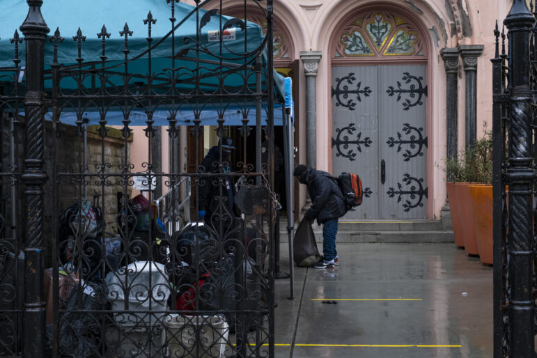 Behind a black iron gate, suitcases, buckets covered in clear plastic, and black trash bags full of items sit under a blue canopy. A man wearing a black Raiders hat faces a person wearing a black mask who carries an orange backpack. The person with the backpack is holding the top of another bag that rests on the ground. In the background, one set of the grey doors of St. Boniface Catholic Church are open, while another is closed.