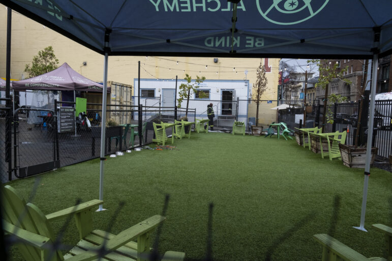 A black canopy that says "Urban Alchemy" in green font stands over a patch of green astroturf. The astroturf rectangle is enclosed by black fencing, and on the inside of the fence 7 lime green wooden recliner chairs are turned sideways. In the far background, a man is walking in front of a while portable building in a black jacket and black pants with neon yellow and reflective stripes.
