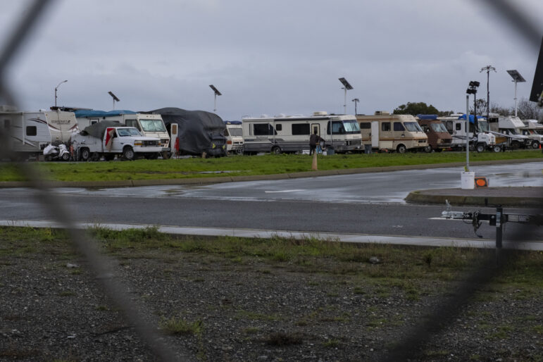 A line of 10 recreational vehicles rests behind a patch of green grass in a parking lot. The pavement in front of the grass is wet, and some puddles of rainwater have gathered in the street.