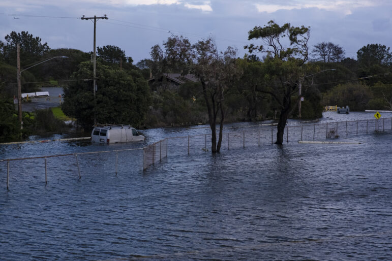 A chain-link fence runs through several feet of water that has accumulated on the street due to flooding. Behind the fence, a white van is partially submerged. Two large trees also stand in the water in front of the fence.