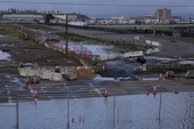 A chain link fence and three orange cones are submerged in several feet of water. Behind the flood, 10 orange cones line a roadway around puddles and flooding to warn passerby. Two woman speak to each other. Several concrete barricades are lined up along the street and around areas of pooled water.