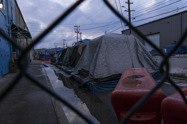Through the wire of a chain-link fence, a line of four tents can be seen resting atop platforms. The tents are covered by tarps in muted colors to keep out rain, though to the left of the tents water has pooled next to a curb. Four plastic orange barricades are placed several feet in front of the first and last tents in the row.