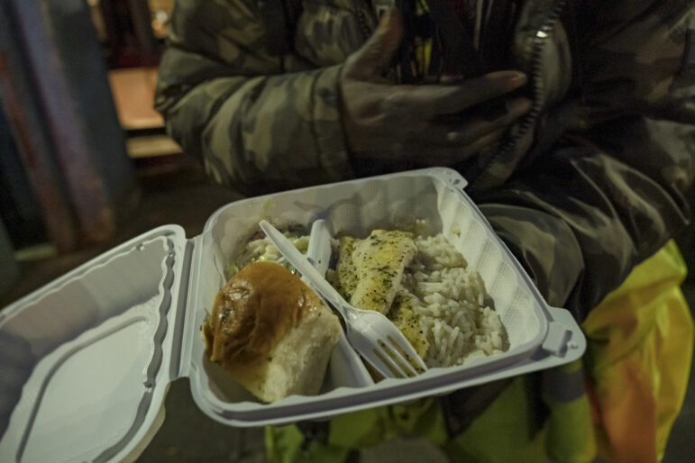 A man in a camoflauge jacket holds in one hand a white take-out container that is opened to reveal a piece of bread and fish sitting atop a bed of white rice. A white plastic fork is laid across the food. The man holds his other hand against his abdomen, and his face is not visible.