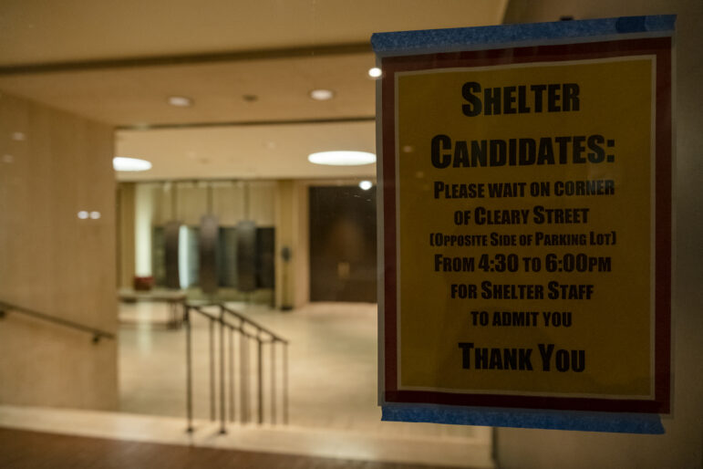 A yellow poster with a red border and black text is stuck to a door with blue masking tape. The poster reads: "Shelter candidates: Please wait on corner of Cleary Street [opposite side of parking lot] from 4:30 to 6:00 pm for shelter staff to admit you
Thank you" in all caps. Behind the glass door inside, a short descending staircase is illuminated by overhead lights.