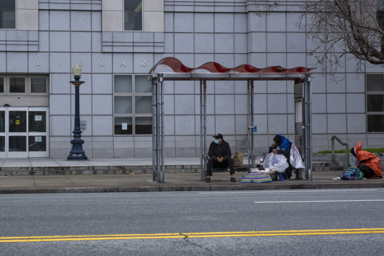 Two men sit underneath the red overhang of a Muni bus stop in front of a grey building. One man is wearing a blue jacket and has a box in front of him that is covered by a colorful striped umbrella. To his left, another person in an orange poncho is squatting on the ground looking at the contents of a multi-colored bag.