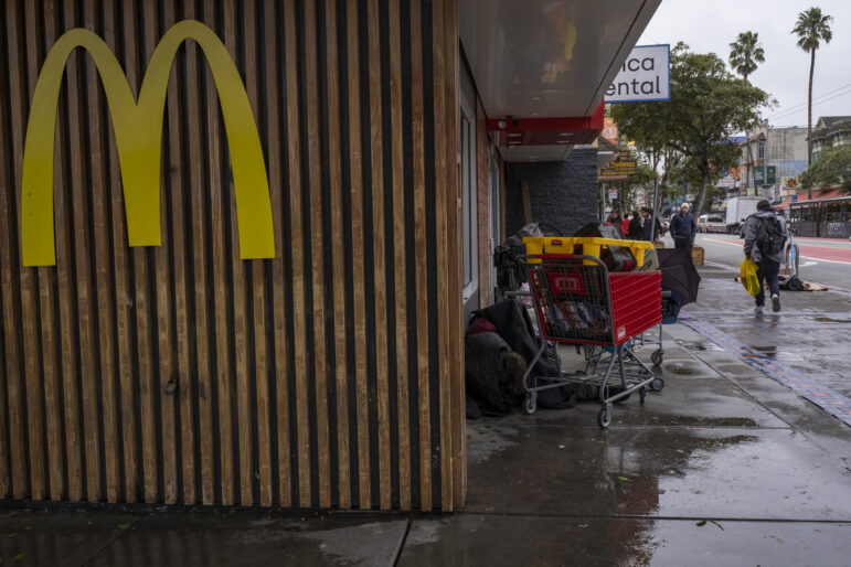 A large yellow M in the McDonald's font hangs on the front of the wood paneling of a storefront. To the right, a man is nestled between the wall of the fast food joint and a red Lowe's shopping cart filled with items. Several suitcases are stacked up behind the man. To the right of the cart and bags, people walk along the sidewalk.