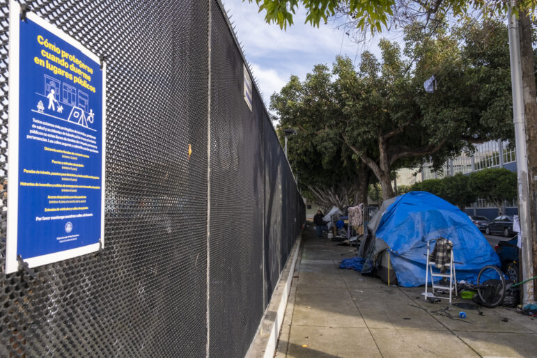 A blue poster with yellow and font is attached to a black chain link fence with black zip ties. The text on the poster is written in Spanish and discourages people from camping. To the right of the fence, a tent covered in blue tarps is surrounded by a step-up ladder, a bike wheel, and other personal items. Behind the fence, several other tents and structures have been erected. A person in a black jacket walks between the fence and the tents.
