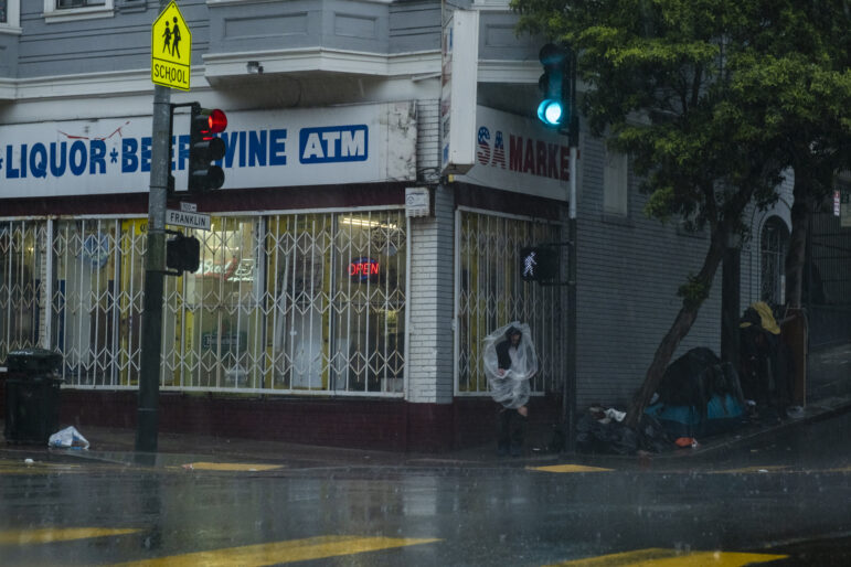 A person in ripped black pants stands in front of a liquor store in a clear plastic bag to shield themselves from the rain. Behind them, a street signal light turns green. Nearby, a tree shields a tent that is surrounded by black trash bags and other personal items.