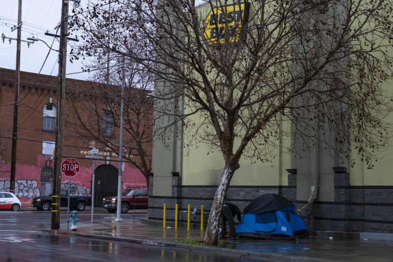 Next to a light yellow building with a large yellow Best Buy logo, a man in all black secures a blue tent under a large tree.
