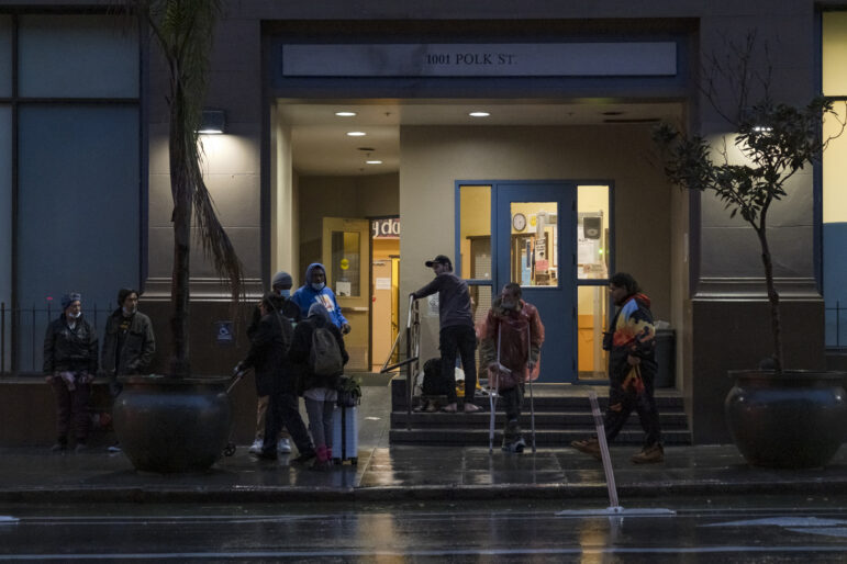 Nine people stand outside the doors of a yellow and blue building with blue doors that is labeled "1001 Polk St." One man in a plastic red poncho is standing on crutches, and another person is wearing a backpack and rolling a blue suitcase behind them. Inside the building, the lights are on but no one is at the door.