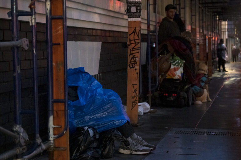 A person sits against part of a brick building, the upper half of their body covered in a blue plastic poncho that reads "St. Anthony's hope served daily" in all caps. Down the sidewalk, a man stands next to his dog who is sitting on the wet pavement.