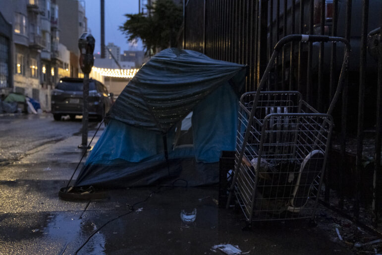 A blue tent is covered by a black rain fly that is attached to a nearby fence. In front of the tent, a folding shopping cart carries a shoe and several other indistinguishable items. A parking meter are car visible near the sidewalk behind the tent.