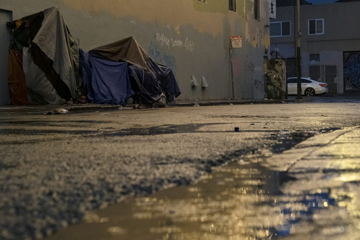 Two makeshift shelter structures are covered in a variety of mutely colored tarps to keep out the rain. They are built along a multi-colored wall that displays some graffiti. A parking sign nearby reads "Tow Away no parking any time" in all caps. In the foreground of the picture, puddles in the street are reflecting light from street lamps.