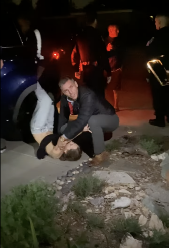 A teenage girl wearing jeans and a black top is struggling with her legs in the air as she is pinned down in a driveway by a man wearing dark clothing next to a large black SUV with rocks and shrub landscaping in the foregound. Police officers are standing in a circle in the background talking to each other, not watching the man and girl.