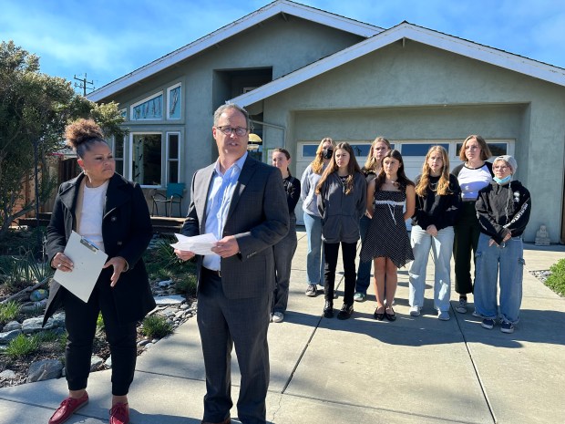 A man and a woman stand in the foreground holding papers as the man speaks during a press conference. A group of people stands behind them in a driveway in front of a beige colored single-family home.