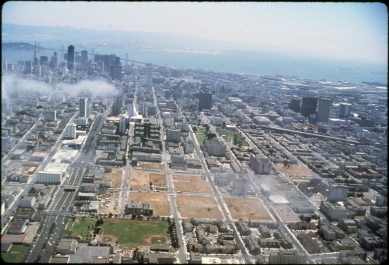 In the forefront, hundreds of buildings, mostly low-rise, surround six empty blocks covered by dead grass in the Western Addition neighborhood. In the top left background, the skyscrapers of downtown and the Bay Bridge are visible.