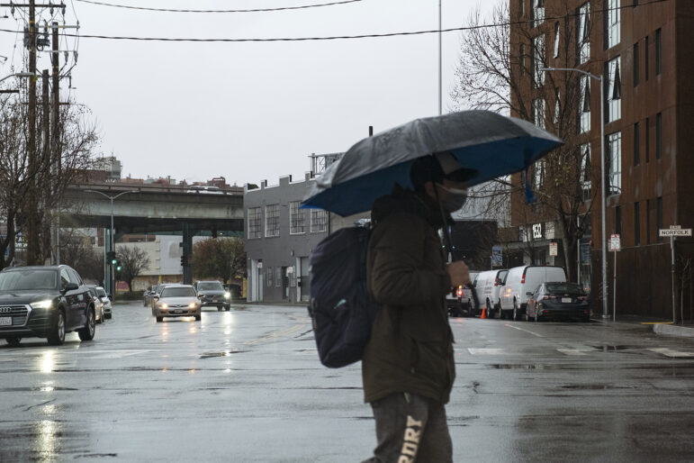 A person wearing dark clothing and a backpack carries a navy umbrella while crossing a city street in the rain. The sky is cloudy and gray. Traffic is light.