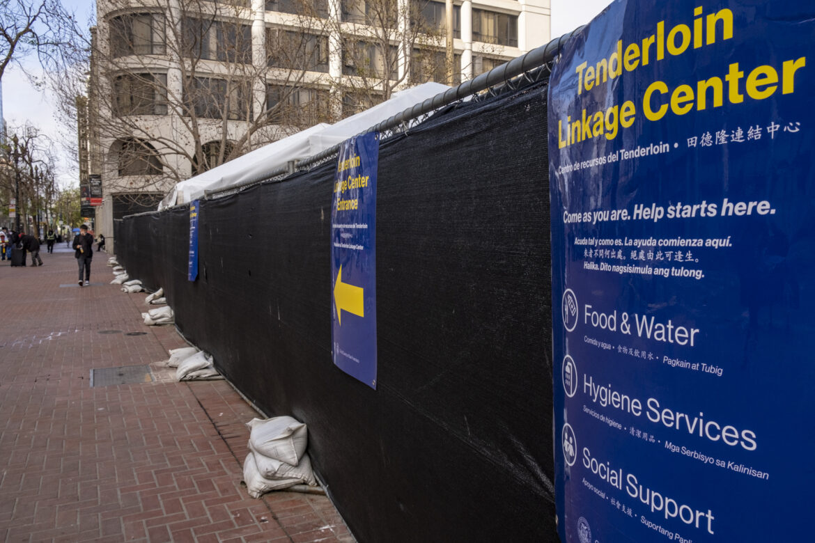 A fence enclosing the Tenderloin Linkage Center at United Nations Plaza in San Francisco is covered in black tarp and signs promoting the center and its services. Pedestrians walk along a red brick-paved sidewalk outside of the enclosed area.