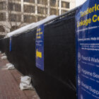 A fence enclosing the Tenderloin Linkage Center at United Nations Plaza in San Francisco is covered in black tarp and signs promoting the center and its services. Pedestrians walk along a red brick-paved sidewalk outside of the enclosed area.