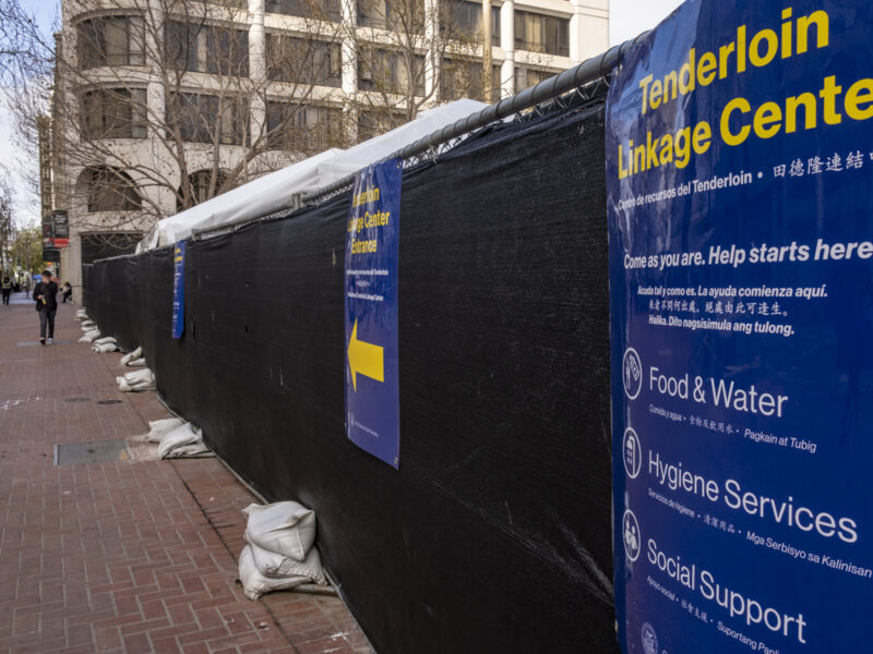 A fence enclosing the Tenderloin Linkage Center at United Nations Plaza in San Francisco is covered in black tarp and signs promoting the center and its services. Pedestrians walk along a red brick-paved sidewalk outside of the enclosed area.